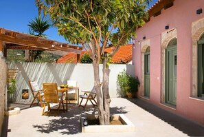 A courtyard with a tree, wooden chairs, a table with fruits, and a building with a pink facade and green doors. - Vineyard View, next to Best Wineries, "Trigos" appartment (52QJ+W6 Avgeniki)