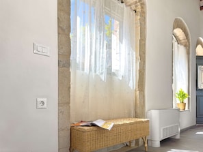 Ein Zimmer mit einer Steinswand, einer Korbsitzbank mit einer Zeitschrift, einem Heizkörper und einem Fenster mit durchsichtigen Vorhängen.