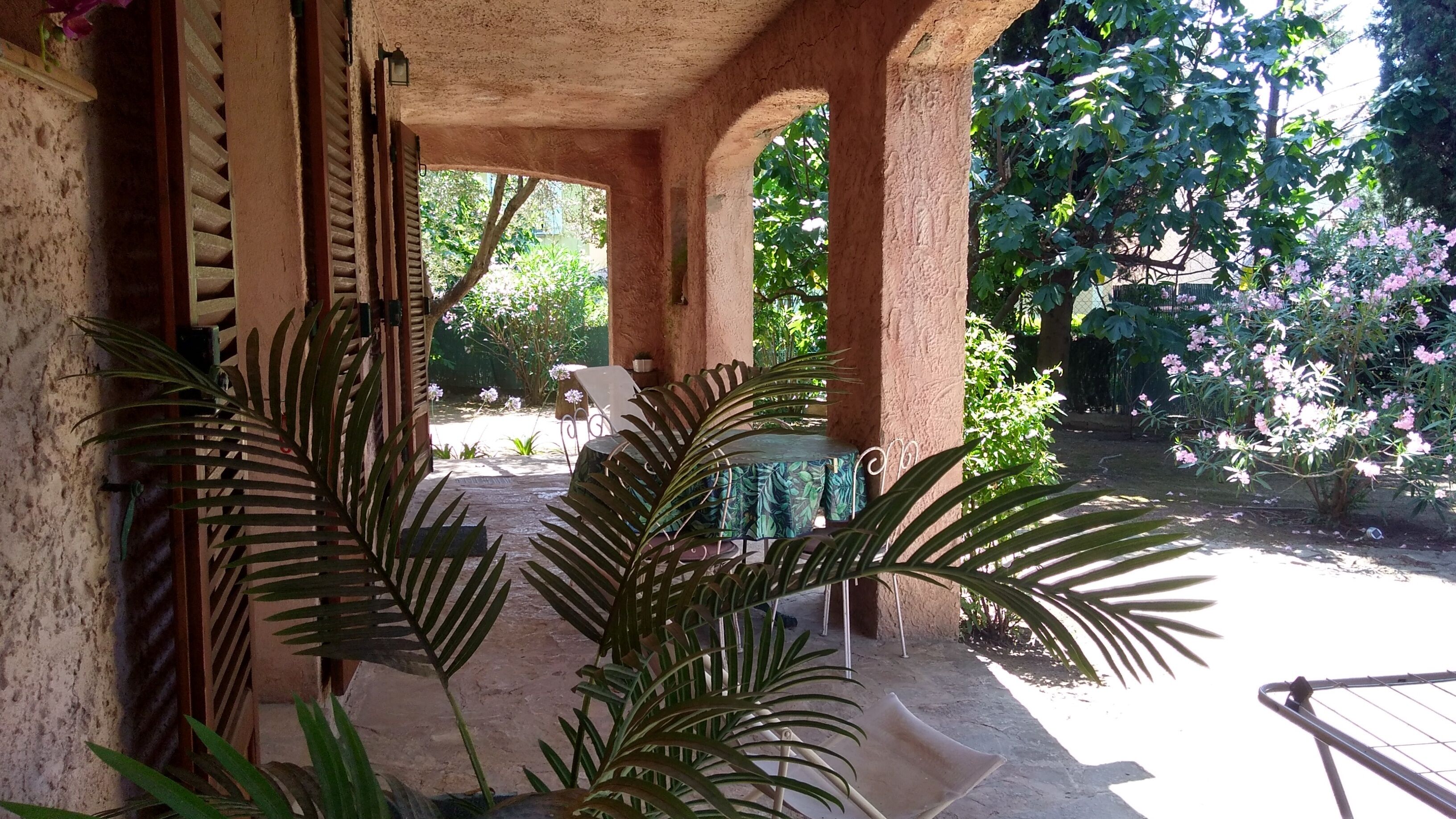 Un espacio al aire libre techado con mesa y sillas, rodeado de abundante follaje y plantas en flor.