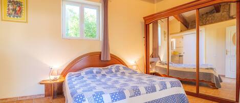 A bedroom with a wooden ceiling, a large window with a curtain, a bed with a patterned bedspread, and a sliding glass door leading to another room.