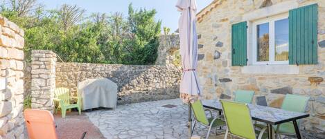 A stone building with a stone wall, a window with green shutters, outdoor seating with chairs and a table, and an umbrella.