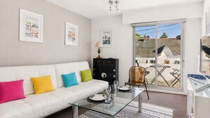 En moderne stue med en hvid sofa, farverige puder, et glasbord, et sort skab og udsigt udad gennem et stort vindue.