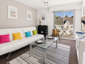 Ein modernes Wohnzimmer mit einem weißen Sofa, bunten Kissen, einem Glastisch, einer schwarzen Kommode und Blick ins Freie durch ein großes Fenster.