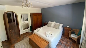 Une chambre à coucher comprenant un lit, une armoire en bois, un miroir, un lustre, une table de chevet et une chaise en osier.