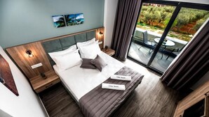 A modern hotel room with a large bed, a nightstand, a bedside table, a window with a view of the outdoors, and a small table with chairs outside. - Calda Resort (Polygyros)