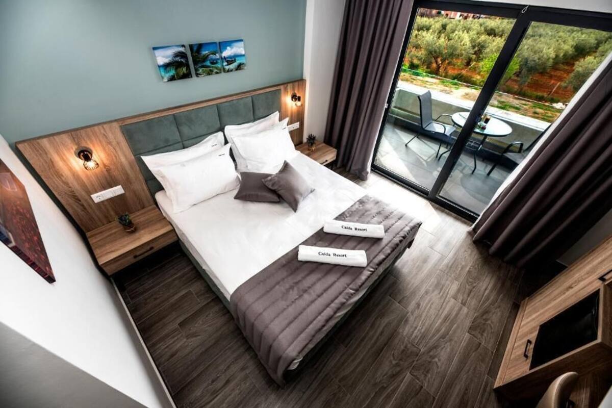 A modern hotel room with a large bed, a nightstand, a bedside table, a window with a view of the outdoors, and a small table with chairs outside.