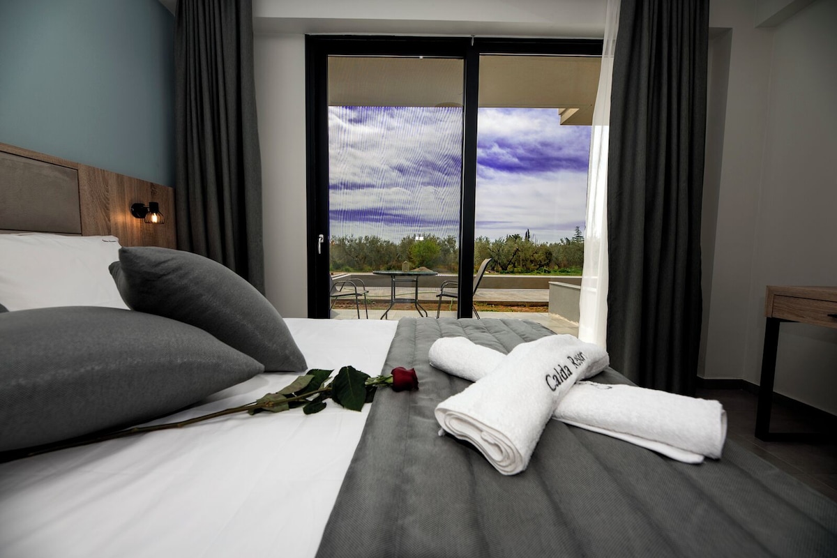 Une chambre d’hôtel avec un lit, des oreillers, une serviette roulée, une rose et une vue sur l’extérieur grâce à une porte coulissante.
