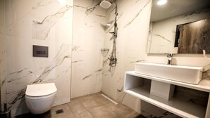 A bathroom with a marble wall, a white toilet, a shower, and a white vanity with a mirror. - Calda Resort (Polygyros)