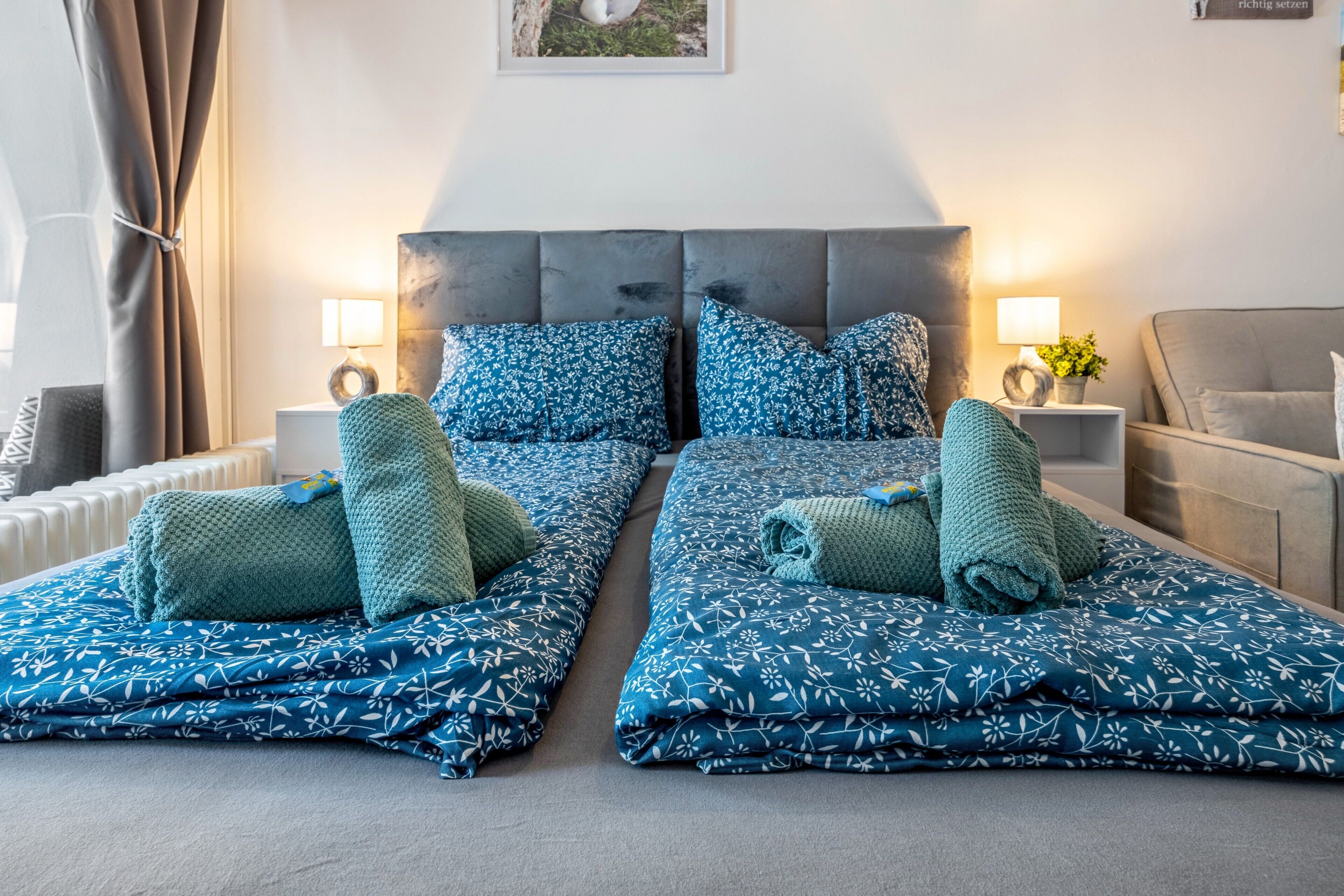 A double bed with matching blue floral bedding, two bedside tables with lamps, a grey sofa, and a wall-mounted picture.