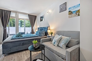 Un salon moderne avec un canapé, une petite table sur laquelle est posée une plante, et un lit recouvert d’une couverture bleue.