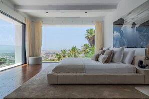 A modern bedroom with a large bed, a view of the city, and a painting on the wall. - Villa Isola Bianca with pool - My Rental Homes (Playa De Talamanca)