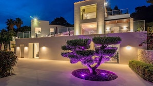 A modern two-story house with a balcony, a well-lit driveway, and a landscaped garden with a tree. - Villa Isola Bianca with pool - My Rental Homes (Playa De Talamanca)