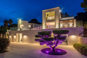 A modern two-story house with a balcony, a well-lit driveway, and a landscaped garden with a tree. - Villa Isola Bianca with pool - My Rental Homes (Playa De Talamanca)