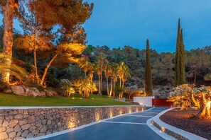 A landscaped pathway with a stone wall, palm trees, and a tall coniferous tree. - Villa Isola Bianca with pool - My Rental Homes (Playa De Talamanca)