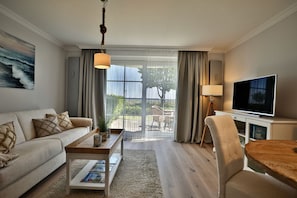 A living room with a sofa, coffee table, TV, and a view through a large window. - Beach shell 1 - Live directly on the beach - dreamlike vacation accommodation with (Scharbeutz)
