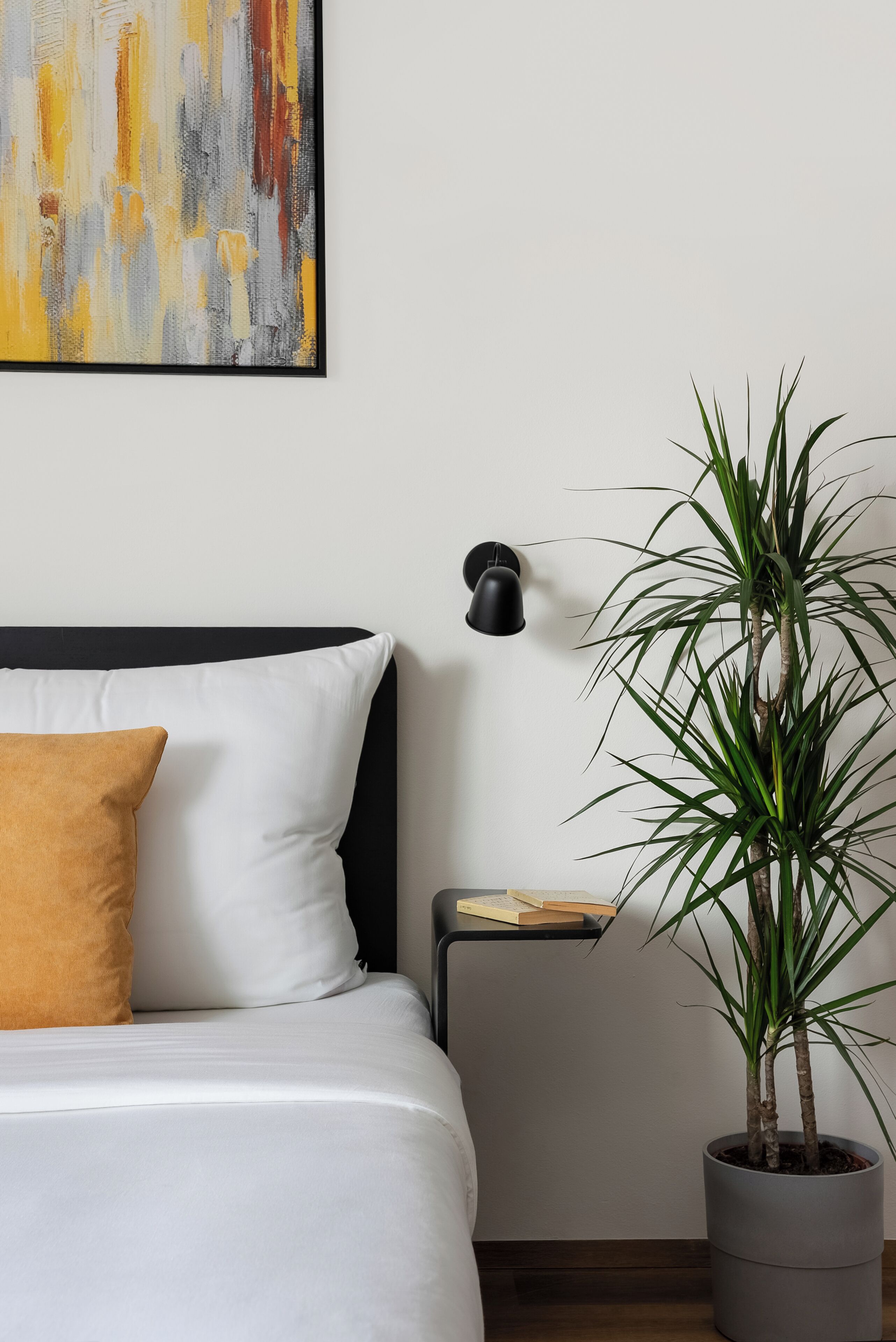 Un lit avec des oreillers blancs et moutarde, une table de chevet avec un livre, une plante en pot et une lampe fixée au mur.