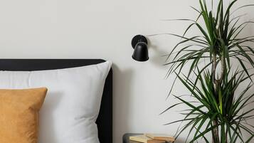 A bed with white and mustard pillows, a bedside table with a book, a potted plant, and a wall-mounted lamp.