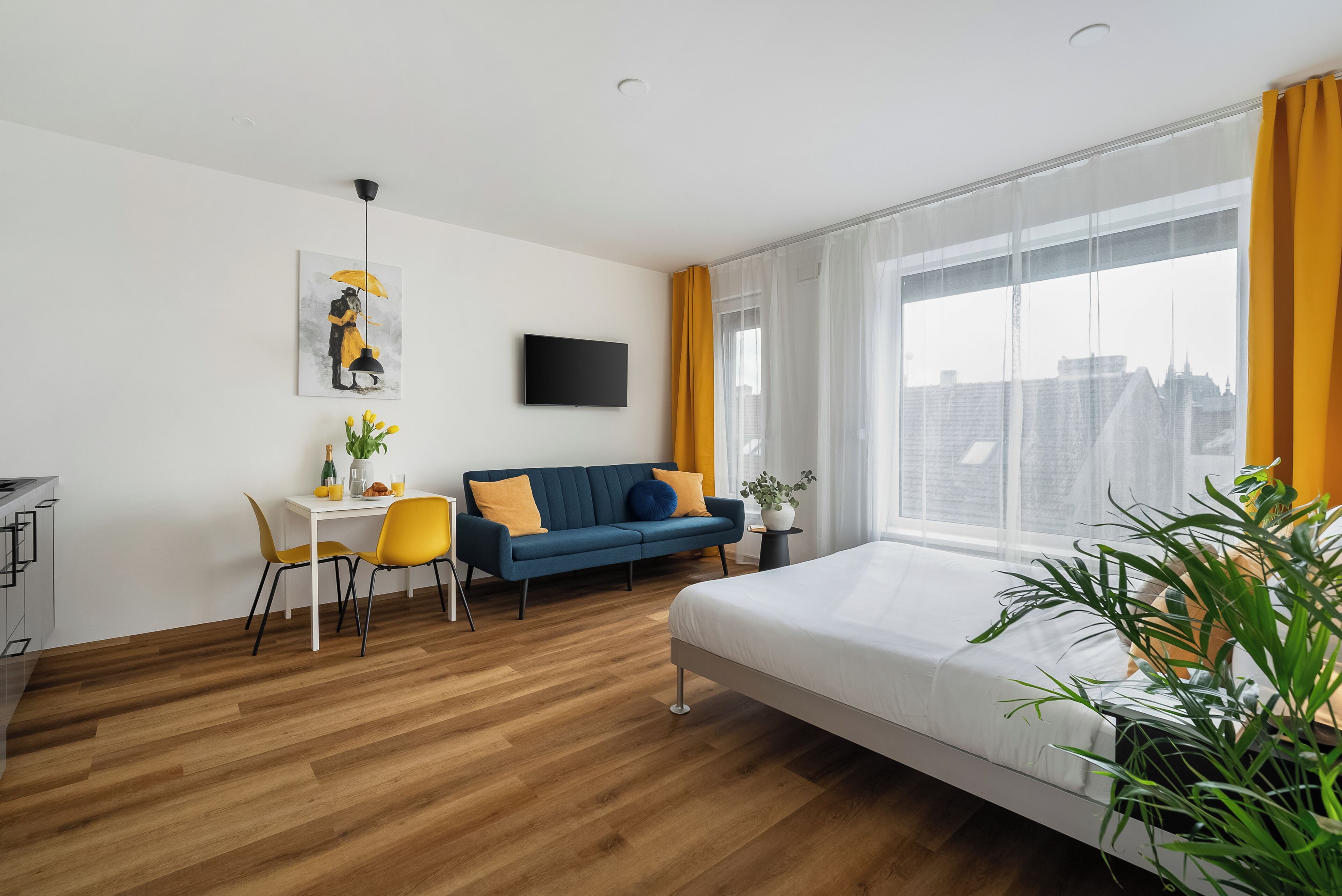 A modern hotel room with a bed, a sofa, a dining table with chairs, a TV, and a large window with curtains.