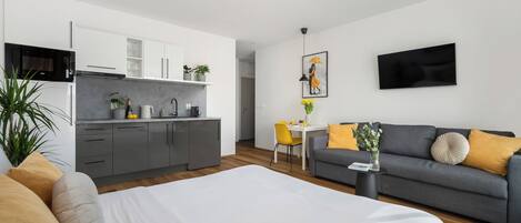 Una camera d'albergo moderna con un letto, un divano grigio, un tavolo da pranzo con sedie gialle, un angolo cottura e una televisione a schermo piatto montata a parete.