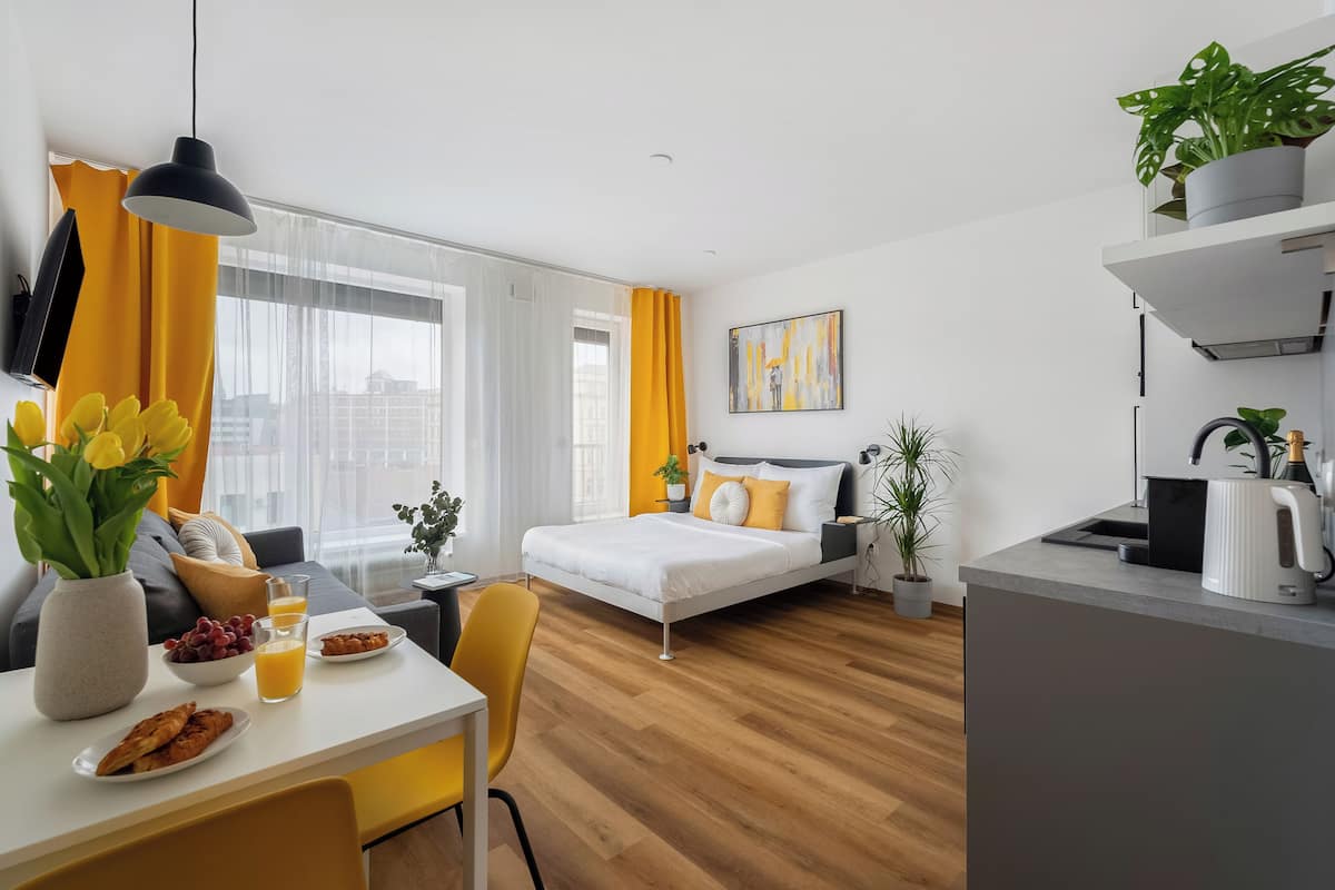 Una habitación de hotel moderna con cama, un área de comedor con desayuno, una cocina y un amplio ventanal con cortinas.