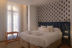 Ein Schlafzimmer mit einem Bett, einem Nachttisch aus Holz, einem kleinen Hocker und einem Fenster mit leichtem Vorhang.