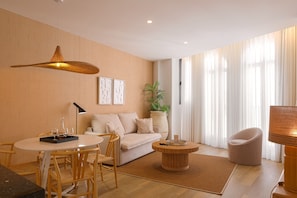 Ein modernes Wohnzimmer mit Sofa, Esstisch, Stühlen, Couchtisch, Stehlampe, Pendelleuchte, einem Blumentopf und einem großen Fenster mit Vorhängen.