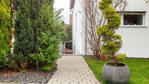 Un chemin pavé mène à un bâtiment à l’extérieur blanc, entouré de différents arbres et d’un grand bac à fleurs.