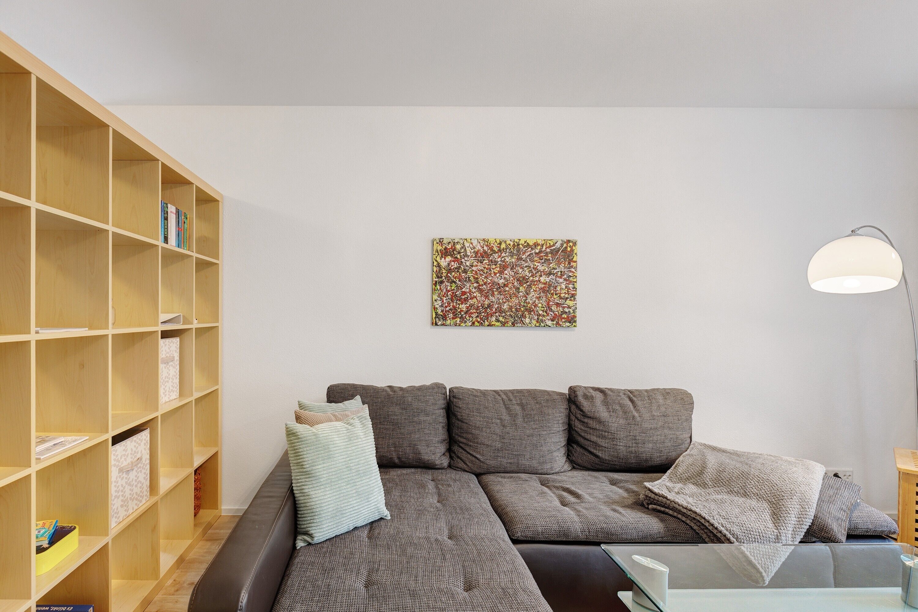 A modern living room with a sectional sofa, a bookshelf, a lamp, and a colorful abstract painting on the wall.