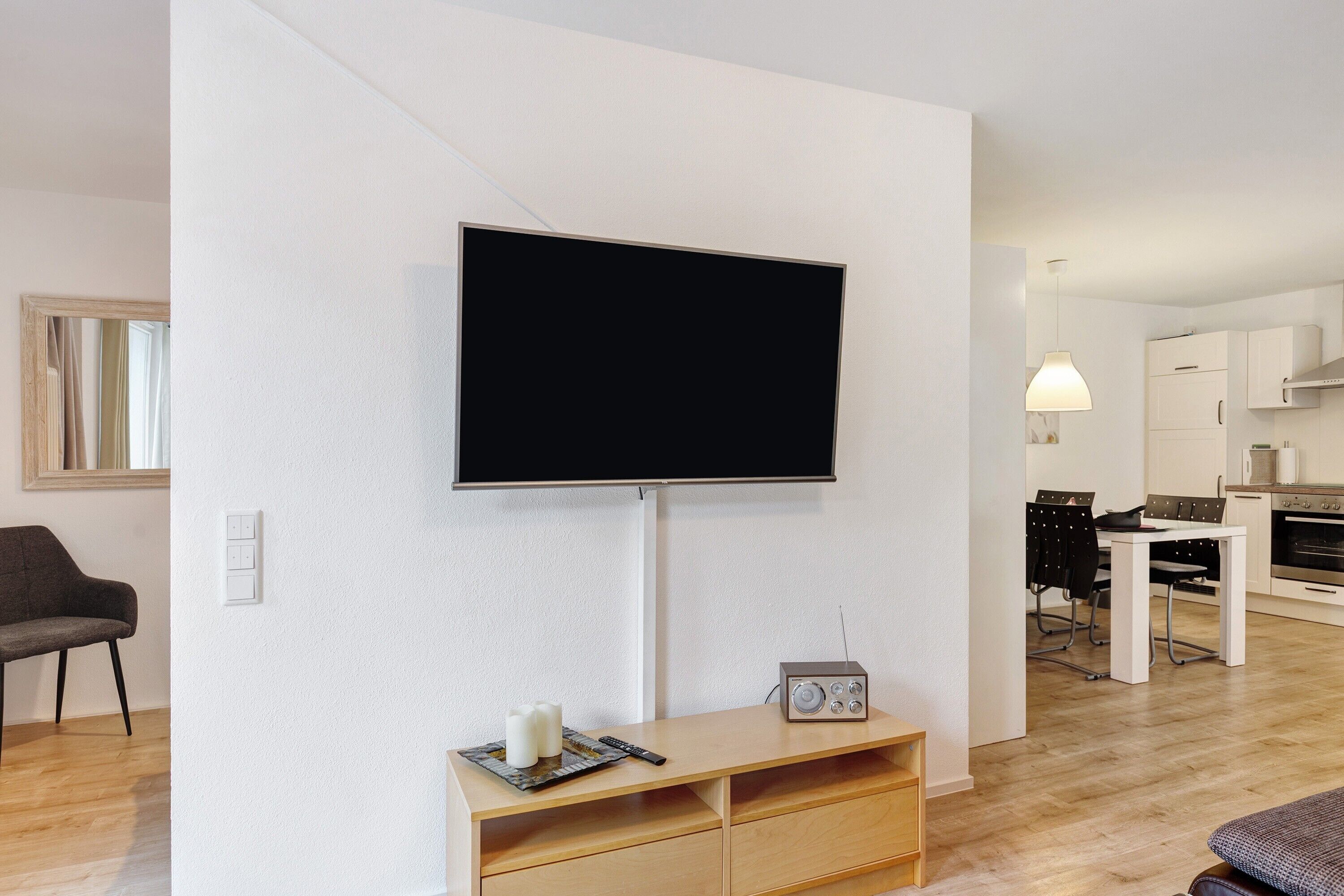 Ein modernes Wohnzimmer mit einem flachbildfernseher an der Wand, einer Holz-Unterhaltungseinrichtung, einem Essbereich mit Tisch und Stühlen sowie einer Küche im Hintergrund.