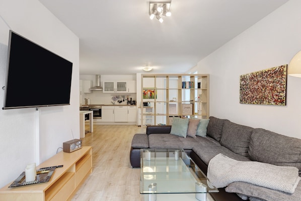 Ein Wohnzimmer mit einer Couch, einem Couchtisch, einem Flachbildfernseher und einer Küche im Hintergrund.