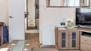 Quarto pequeno com uma cama, uma mesa com cartas, um radiador, um armário com uma TV e uma porta que dá para outro quarto.