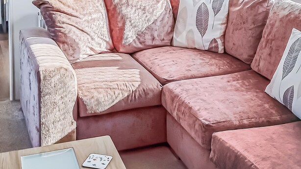 Um sofá seccional rosa com almofadas decorativas com franjas e um jogo de cartas na lateral.