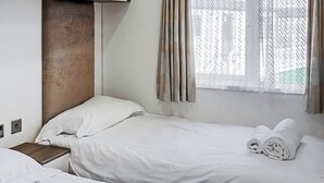 Quarto de hotel com duas camas, cabeceira de madeira, janela com cortinas e uma mesinha de cabeceira.