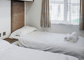 Ein Hotelzimmer mit zwei Betten, einem Holzkopfende, einem Fenster mit Vorhängen und einem kleinen Nachttisch.