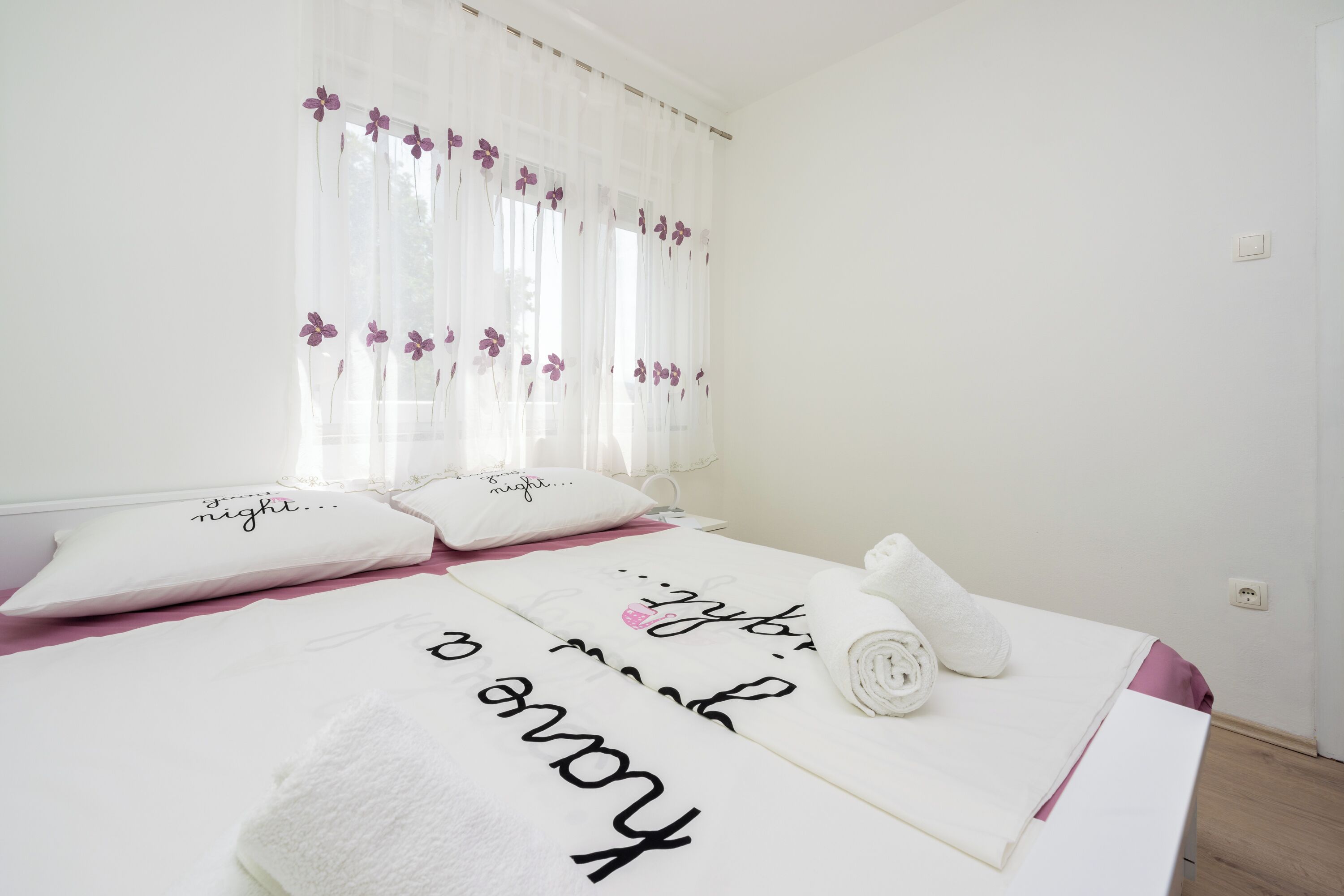 Una cama bien hecha con una toalla enrollada y almohadas, una pared blanca y una ventana con cortinas traslúcidas.