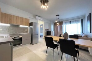 Une cuisine moderne dotée d’un four et d’un réfrigérateur encastrés, un coin repas avec une table en bois et des chaises noires, et un salon avec un canapé et une grande fenêtre.