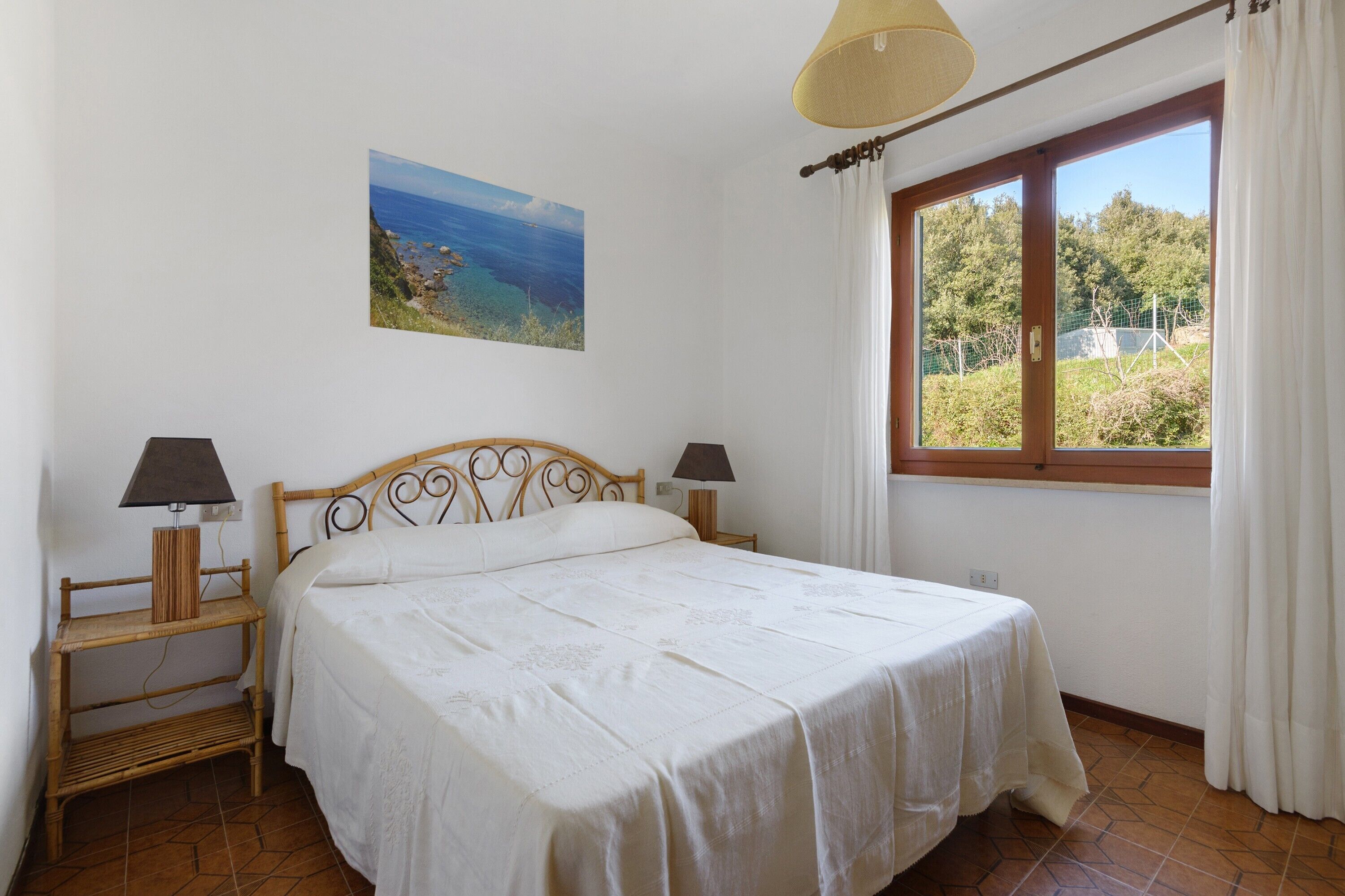 Un dormitorio con cama, dos mesitas de noche, lámpara, ventana con cortinas y un cuadro en la pared.