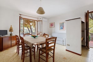 Un comedor con una mesa y sillas de madera, un televisor sobre un mueble, y una ventana con vistas a árboles.