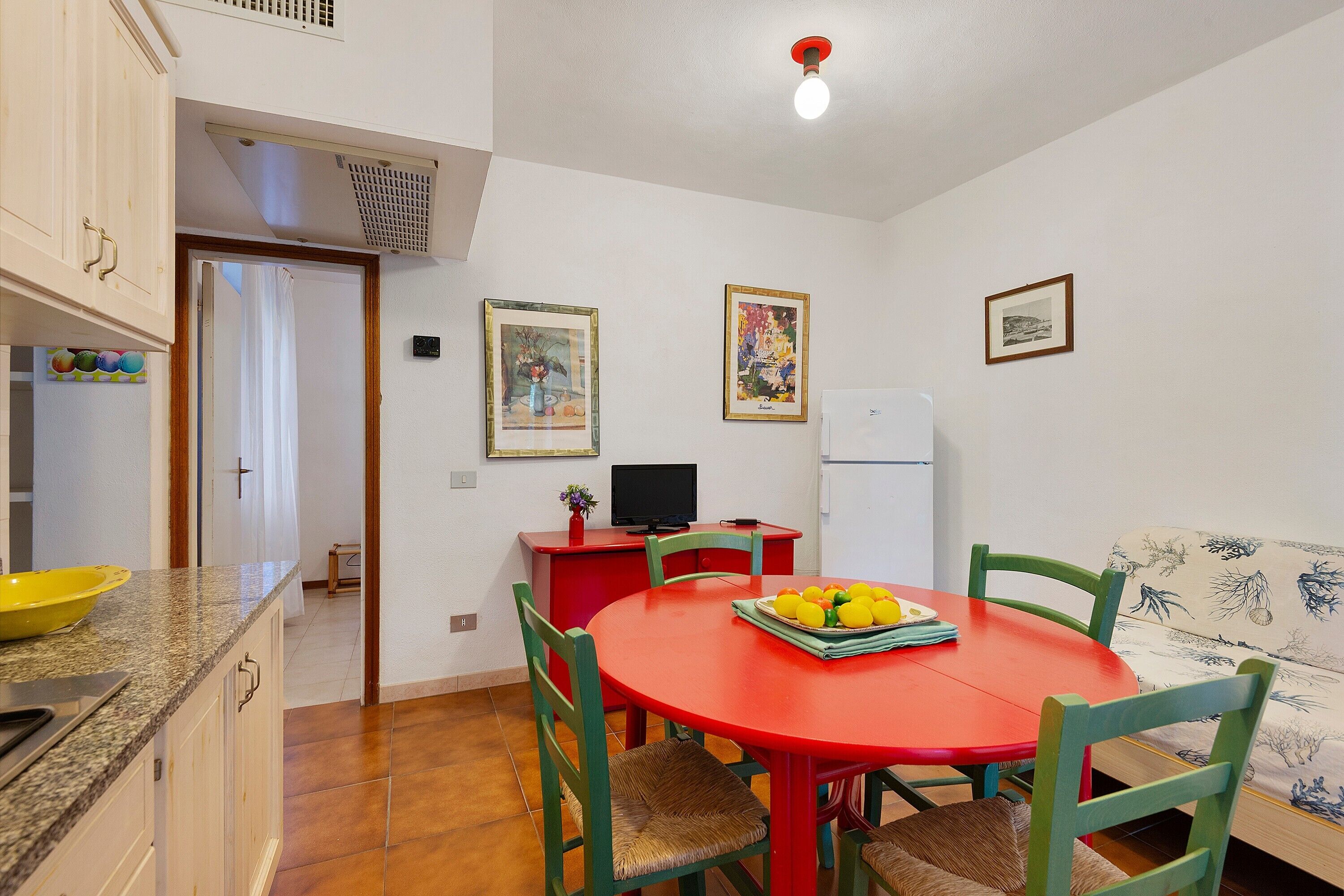 Une cuisine compacte avec une table à manger rouge, des chaises vertes, un saladier jaune et un petit téléviseur.