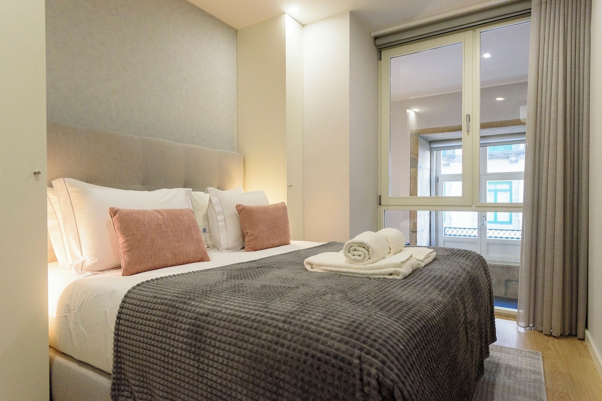 A hotel room with a bed, pillows, a blanket, a window with curtains, and a view of the outside.