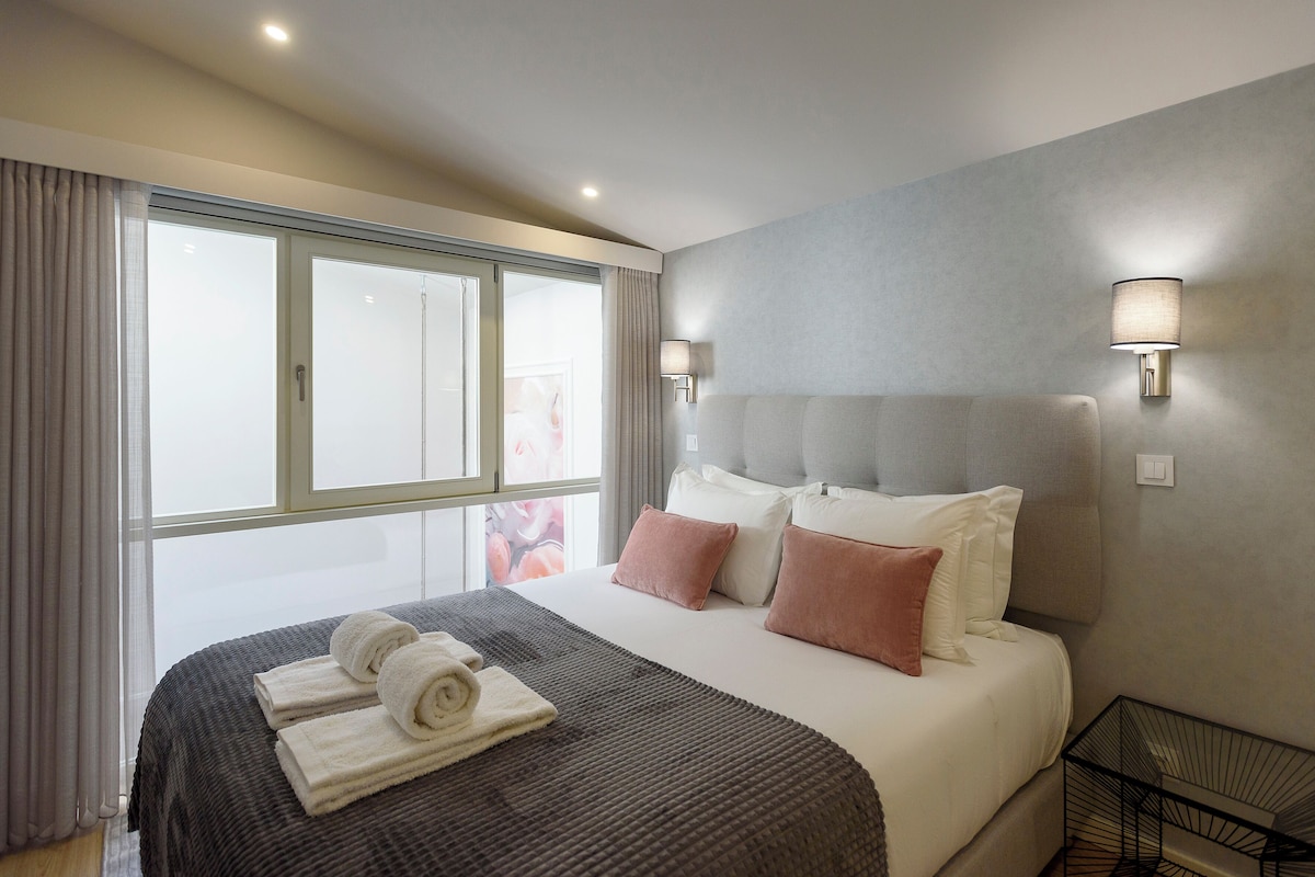 A hotel room with a large bed, two wall-mounted lamps, a window with curtains, and a bedside table with a lamp.