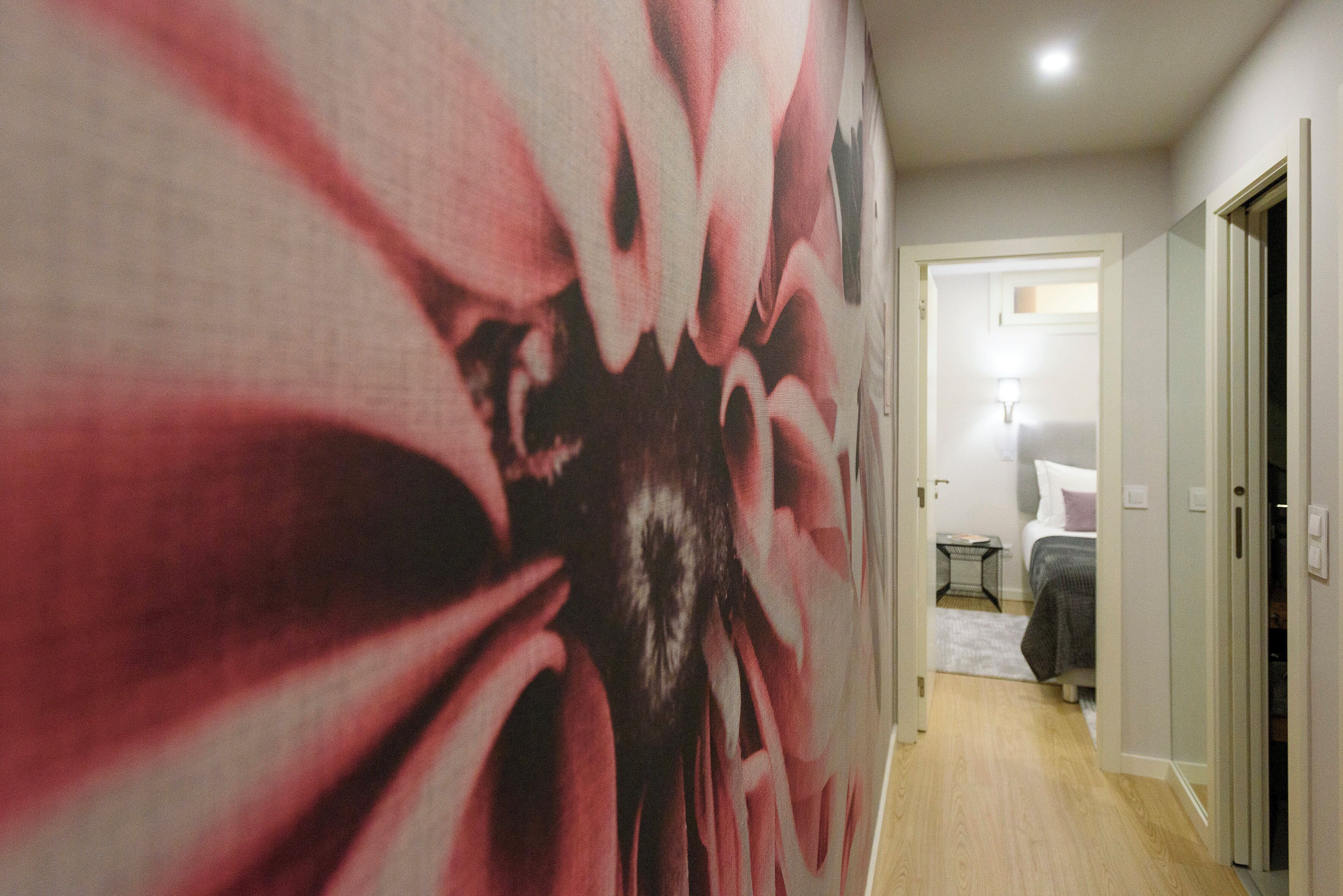 A hallway with a large floral mural, a bed with a white and gray bedspread, and a small table with a lamp.