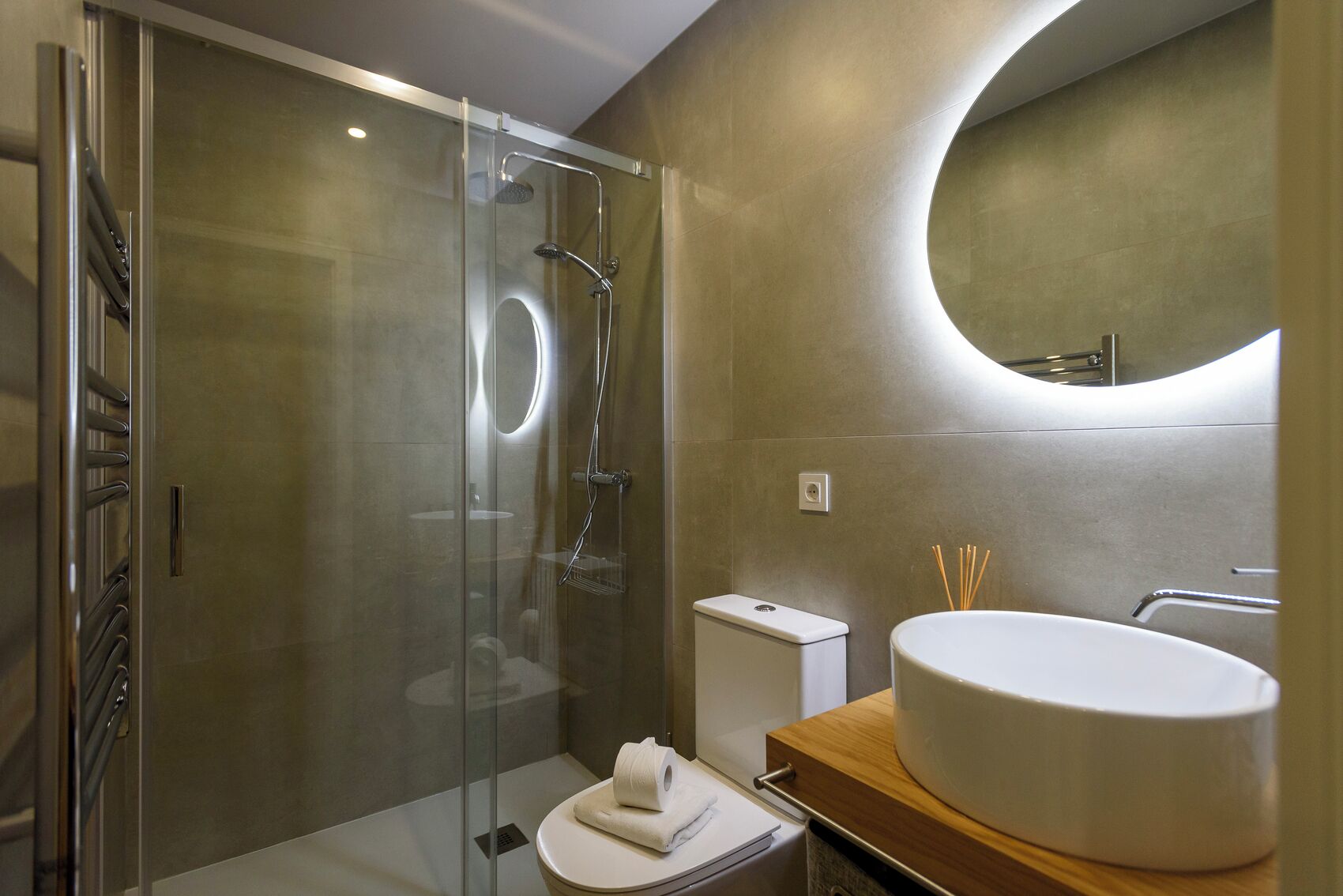 A modern bathroom with a glass-enclosed shower, a toilet, a sink with a round basin, and a mirror with a circular design on the wall.