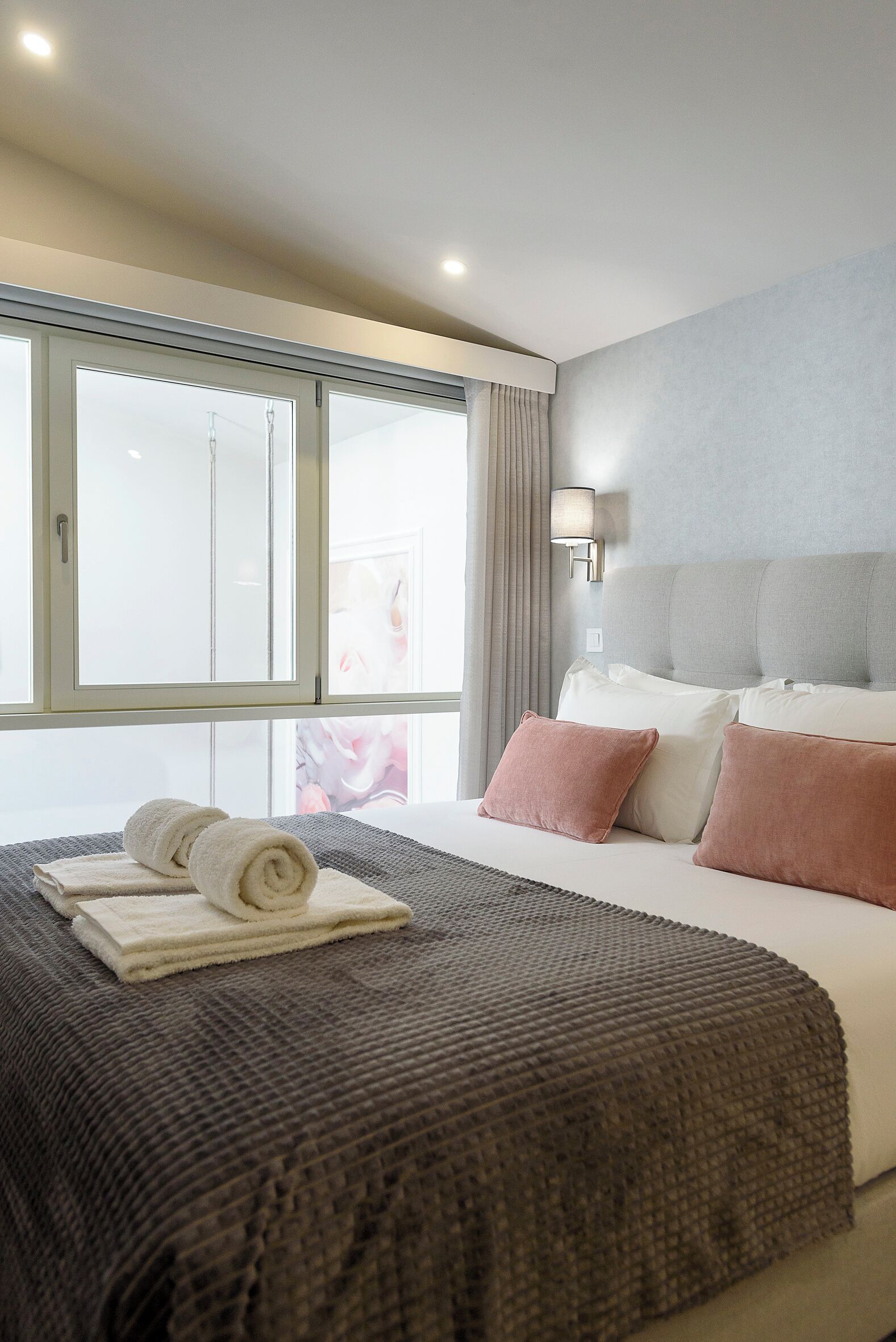 A hotel room with a large bed, a window with curtains, a wall-mounted lamp, and a view of a building outside.