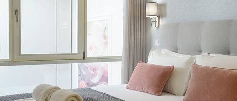 A hotel room with a large bed, a window with curtains, a wall-mounted lamp, and a view of a building outside.