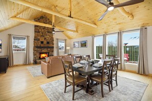 A cozy living room with a stone fireplace, wooden ceiling beams, a dining table with chairs, and large windows with curtains. - A newer 3-bed, 2-bath Pagosa Springs home with mountain views and a 2-car garage. Central Whole House Air Conditioning. Spacious, fully fenced yard — perfect for families, friends, and pets. Newly added hot tub! (Pagosa Springs)
