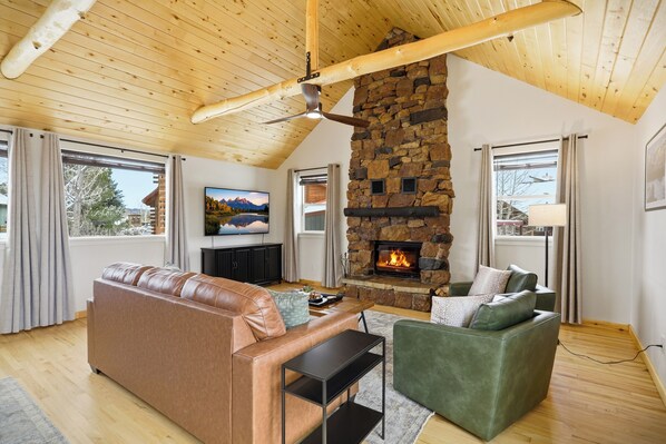 Una sala de estar acogedora con chimenea de piedra, sofás de cuero, ventilador de techo y ventanas grandes.