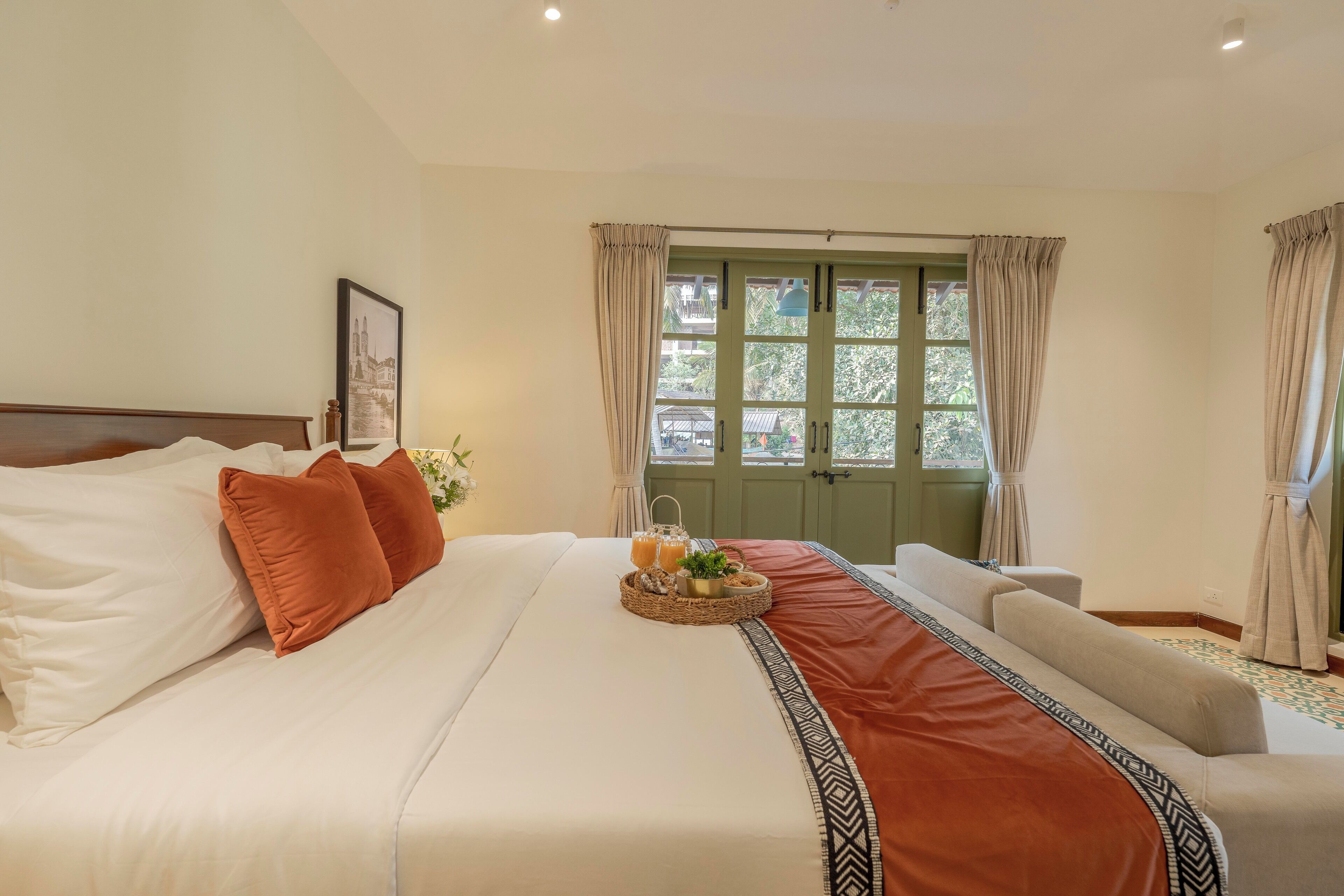 A neatly made bed with pillows, a tray with drinks and a plant, and a view of a green door and windows.