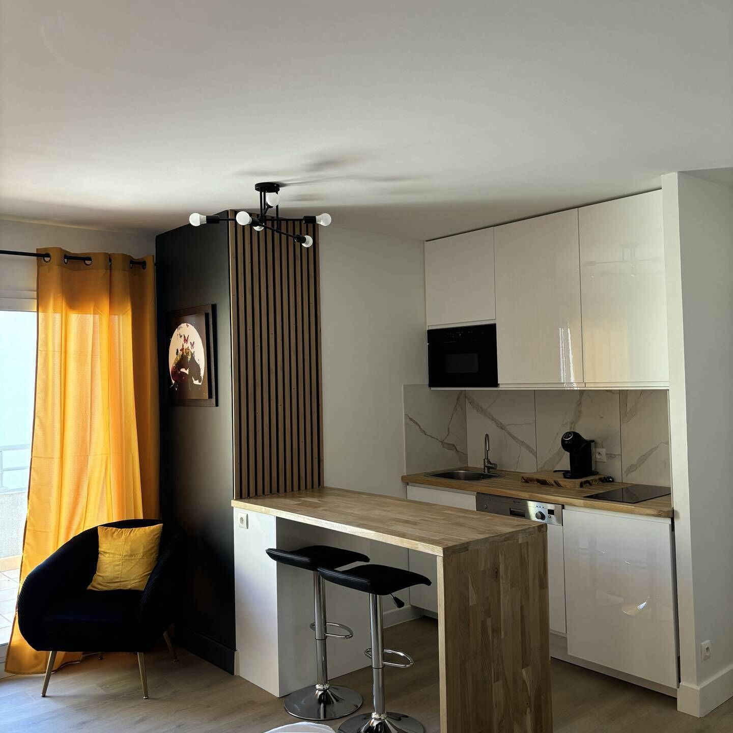 Una cocina moderna con encimera de madera, microondas integrado y lavavajillas. Hay un taburete de bar y una silla con cojín.