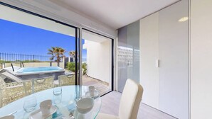 A modern room with a glass table, white chairs, a view of a pool and palm trees, and a sliding glass door. - NAUTILIA  103 - Premium Luxe - Jacuzzi - 2 people (AGDE)